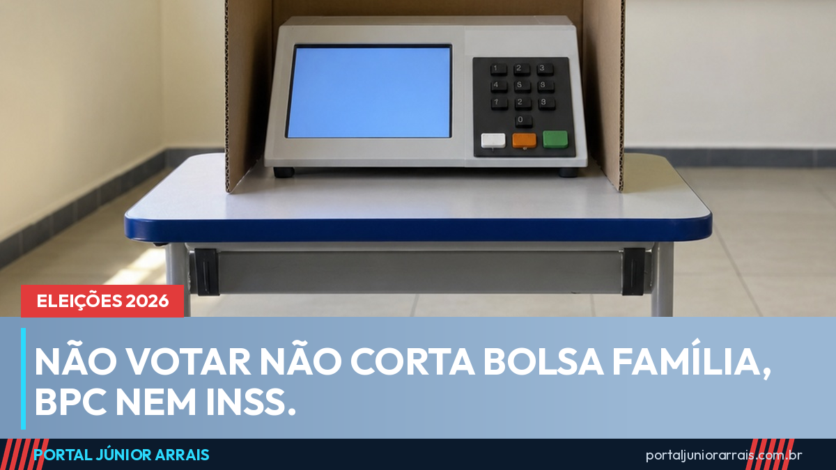 Urna eletrônica ligada dentro de uma cabine de votação de papelão, sobre a carteira de uma sala de aula
